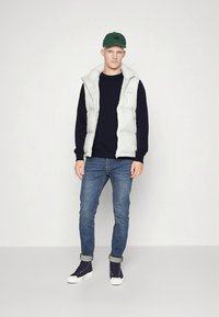 Biela prešívaná vesta cez tmavý sveter, v kombinácii s modrými džínsami a námornícky modrými vysokými teniskami. Doplnky zahŕňajú zelenú čiapku. Jednoduchý dizajn.