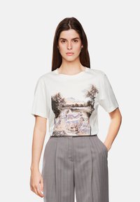 Witte T-shirt met korte mouwen met een kleurrijke pastorale afdruk van koeien en bomen op de voorkant, gecombineerd met grijze high-waisted broeken.