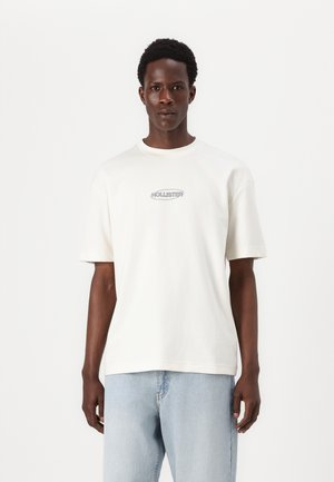 Kremfarget bomull t-skjorte med rund hals, korte ermer og en sentral grå "HOLLISTER" logo. Brukt med lyseblå jeans.