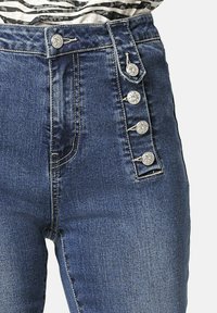Jeans de mezclilla azul de cintura alta con cuatro botones decorativos plateados a lo largo del lado derecho, combinados con una blusa de rayas en blanco y negro.