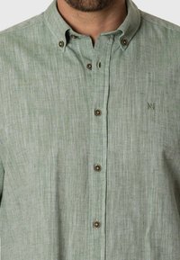 Camisa de lino verde claro con cuello abotonado, que presenta botones marrones y una textura vertical sutil. Emblema bordado en el pecho.