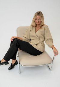 Beige Wickelpullover mit einem Gürtel aus dem gleichen Stoff, strukturiertes Material, ¾-lange Ärmel, kombiniert mit schwarzen, taillierten Hosen und glänzenden schwarzen Schuhen, sitzt auf einem cremefarbenen Stuhl.