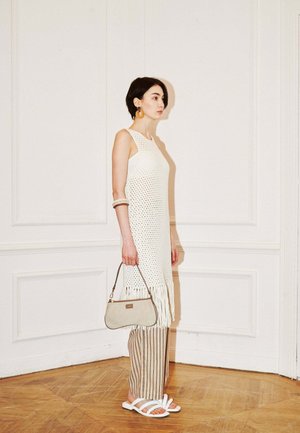 Femme en robe en maille blanche sans manches avec des franges, pantalon rayé, sandales blanches, tenant un sac à main beige contre un mur blanc à panneaux.