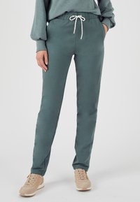 Sweatpants van teal stof, taps toelopend ontwerp, elastische tailleband met witte trekkoord, zijzakken, gecombineerd met beige sneakers.