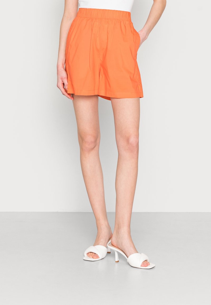 Shorts orange avec une taille élastique, des poches latérales et une coupe décontractée. Portés avec des sandales à talons blancs sur un fond gris.