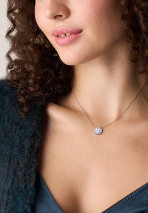 Kroeshaar vrouw die een delicate ronde hangersieraad met sterontwerpen draagt op een dunne ketting, boven een blauwe top en een donkere cardigan.