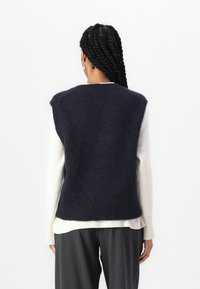Sweater sem mangas em malha azul-marinho com acabamento texturizado e bainha canelada, sobre uma blusa de manga longa creme, combinado com calças cinzentas.