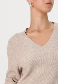 Beige V nyakú pulóver bordázott textúrával. Széles nyakkivágással és laza fazonnal rendelkezik, amit egy gyémánt alakú fülbevalóval párosítottak.