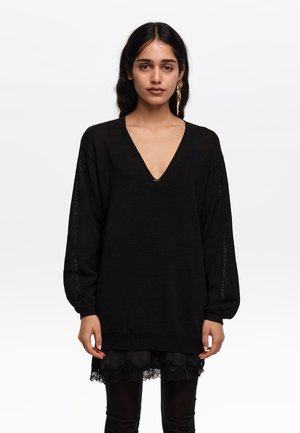 Mujer con cabello oscuro que lleva un suéter negro holgado de cuello en V con mangas abullonadas y un vestido negro con encaje en el dobladillo debajo, sobre fondo liso.