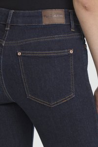 Detailní záběr tmavě modrých džínů s viditelným zadním kapsou, žlutým prošíváním, měděnými nýty a hnědou koženou nášivkou s nápisem "PULZ JEANS".