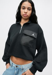 Γυναίκα με μαύρα μακριά μαλλιά, φορώντας μια μαύρη κοντή ζακέτα με φερμουάρ και μεγάλη τσέπη, και ανοιχτό μπλε τζιν, με τα χέρια στις τσέπες.