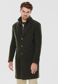 Ciemnozielony wełniany płaszcz z wyłamią kołnierza, trzema guzikami, przednimi kieszeniami i wewnętrzną pikowaną warstwą. Gładka tekstura i dopasowany fason.