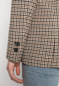 Beżowy, czarny i brązowy żakiet w wzór houndstooth o miękkiej strukturze; posiada czarne guziki na mankietach oraz dopasowany krój.