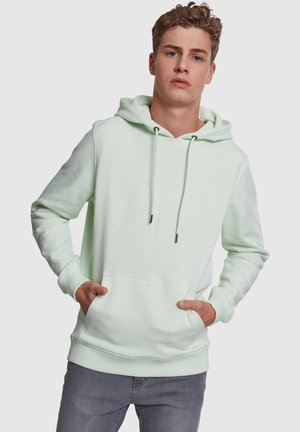 Young man with curly hair wearing a light green hooded sweatshirt with pockets and gray jeans, standing with hands in pockets.