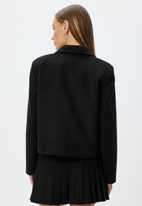 Zwarte wollen mix jas met een gekarteld kraag en een cropped ontwerp, gecombineerd met een geplooide zwarte rok. Effen textuur, minimale details.