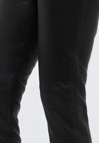 Κοντινό πλάνο μαύρου δερμάτινου παντελονιού που εμφανίζει ομαλή υφή και λεπτομέρειες ραφής στα πόδια πάνω σε απλό φόντο.