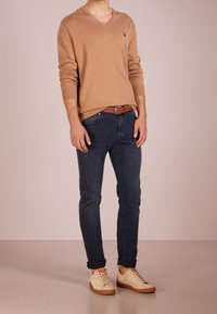 Man som bär en tan V-ringad tröja, mörka uppkavlade jeans med ett flätat brunt bälte och beige sneakers mot en neutral bakgrund.