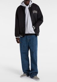 Chaqueta bomber de satén negra con rayas blancas, sudadera gris con capucha, jeans boyfriend azules con patrones intrincados y zapatillas de cuadros sin cordones.