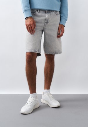 Άνδρας που φοράει ανοιχτό γκρι τζιν σορτς, λευκά αθλητικά παπούτσια με κάλτσες και μπλε μπλουζάκι με μακριά μανίκια, στέκεται μπροστά σε απλή φόντο.