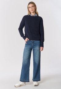 Maglione a maglia blu navy con maniche lunghe, abbinato a jeans a gamba larga blu e sneaker bianche, con un colletto a righe a contrasto sotto.