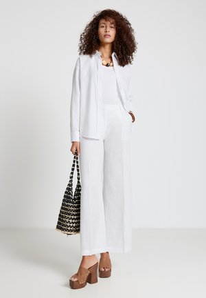 Donna con capelli ricci che indossa una camicia bianca, una canottiera, pantaloni a gamba larga, tacchi platform color cuoio e tiene in mano una borsa tote nera intrecciata.