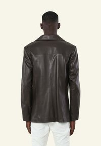 Bruine leren blazer met een geplooide kraag, een zachte textuur en een enkele rugnaad. Gepresenteerd op een model dat witte broeken draagt.