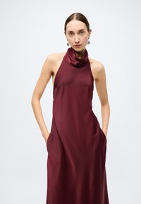 Robe bordeaux en satin à col bénitier et dos nu, avec poches latérales, une texture lisse et une longueur jusqu'au sol.