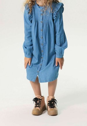 Dziecko w długim, niebieskim sukienkowym fartuszku z guzikami i falbankami na ramionach, stojące w beżowych butach z czarnymi sznurówkami na białym tle.