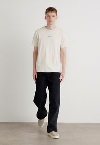 Beige bomull t-skjorte med korte ermer og en liten logo på brystet, kombinert med løse sorte bukser og lyse joggesko.