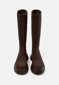 Botas de tela de color marrón oscuro y alto, con suelas de goma gruesa y acanalada, de pie sobre un fondo gris claro liso.