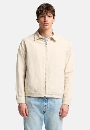 Beige jakke lavet af bomuld med krave, lynlås foran og to sidelommer, kombineret med lyseblå jeans.