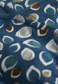 Tessuto blu con un motivo astratto di foglie in bianco, verde acqua, marrone e grigio chiaro. Il tessuto ha una consistenza liscia con una leggera lucentezza.