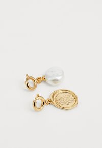 Guld hoops øreringe med en hvid perle på den ene og en guldmedaillon med et tekstureret ugle-design på den anden, på en hvid baggrund.