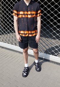 Zwart shirt met oranje-wit patroon, gecombineerd met zwarte shorts. Zwarte sandalen met verstelbare riempjes en witte sokken. Getextureerde achtergrond.