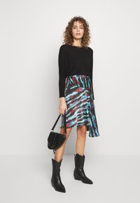 Černý top s dlouhým rukávem, sukně s celobarevným tie-dye vzorem a zavinovacím designem, černé kovbojské boty a malá černá crossbody taška.