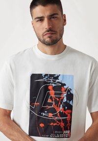 Camiseta blanca de algodón con una impresión gráfica en colores negro, azul y rojo, que incluye una gran 'G' estilizada y patrones florales abstractos.
