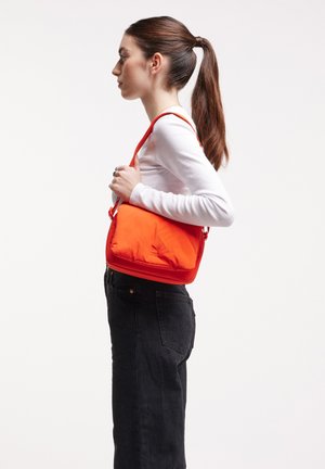 Bolso bandolera de color naranja brillante con textura acolchada, cierre de cremallera y correa ajustable, combinado con jeans negros y una blusa de manga larga blanca.