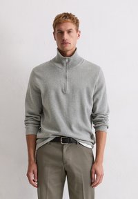 Jasnoszary pulower na zamek wykonany z miękkiego materiału, z wysokim kołnierzem i długimi rękawami, zestawiony z oliwkowozielonymi spodniami i czarnym paskiem.