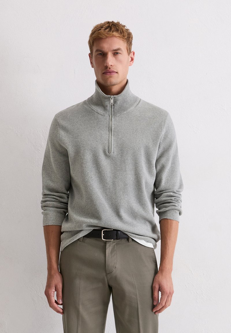 Jasnoszary pulower na zamek wykonany z miękkiego materiału, z wysokim kołnierzem i długimi rękawami, zestawiony z oliwkowozielonymi spodniami i czarnym paskiem.