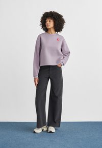 Ανοιχτό μωβ κοντό φούτερ με κόκκινο λογότυπο, σε συνδυασμό με σκούρο παντελόνι φαρδύ και λευκά αθλητικά παπούτσια πάνω σε μπλε πάτωμα.