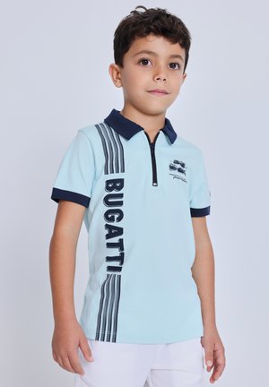 Ragazzo giovane che indossa una polo azzurro chiaro con la scritta "BUGATTI" e colletto blu navy, in piedi davanti a uno sfondo chiaro e uniforme.