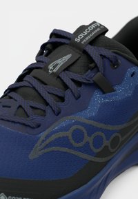 Fialové a čierne trailové bežecké topánky so sieťovaným vrchom s textúrou, výrazným logom a posilneným systémom šnurovania. Obsahujú vzorovanú vrstvu a technológiu Gore-Tex.