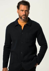 Giacca camicia nera in lana con colletto, chiusura frontale con bottoni e due tasche a toppa. Indossata sopra una t-shirt senape. Texture liscia.