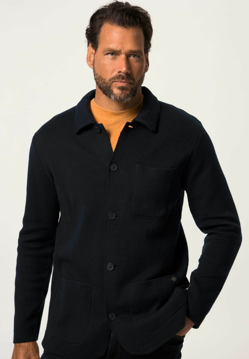 Giacca camicia nera in lana con colletto, chiusura frontale con bottoni e due tasche a toppa. Indossata sopra una t-shirt senape. Texture liscia.