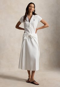 Nő fehér, rövid ujjú polóruhában, elöl csomós díszítéssel, kezét csípőjén tartva, fekete szandált viselve, semleges háttér előtt áll.