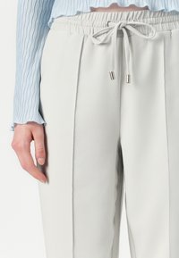 Pantalons de jogging gris clair avec une taille élastique, un cordon de serrage et des accents en métal argenté. La texture semble douce et lisse.