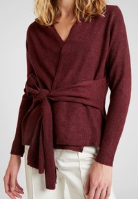 Femme portant un pull cache-cœur bordeaux noué en un nœud à la taille, associé à un pantalon blanc, vue du cou aux hanches.