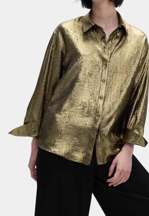 Persoon die een glanzend metallic gouden overhemd met knopen en lange mouwen draagt, gecombineerd met losse zwarte broek tegen een effen lichte achtergrond.