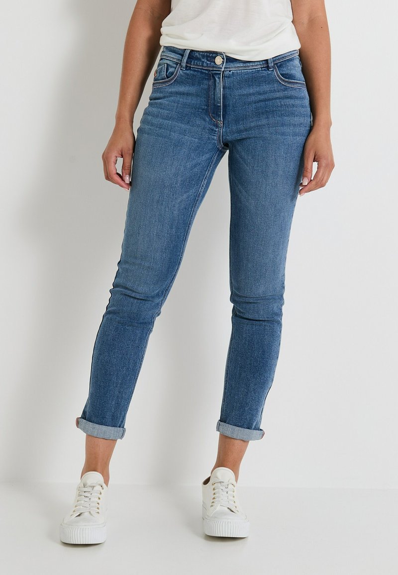 Jeans de denim azul con corte Slim, con cierre de botón, cinco bolsillos y puños enrollados. Combinados con zapatillas blancas.