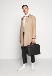 Beige trenchcoat over en hvid skjorte, sorte bukser og hvide sneakers. Holder en sort lædermappe med korte håndtag.